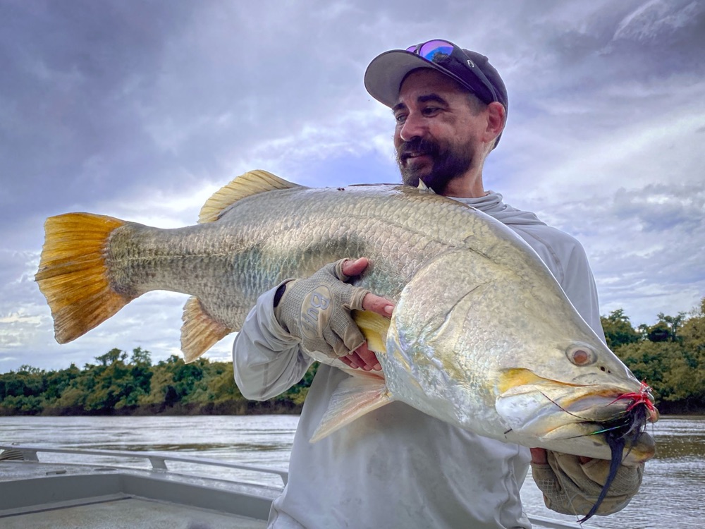 Angler holding a metre-plus barramundi with a bright orange tail under a moody wet-season sky