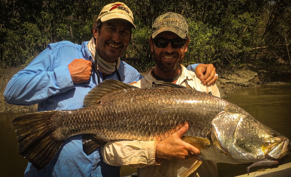 Two grinning anglers holding a big dark-scaled barramundi caught tight to the mangroves